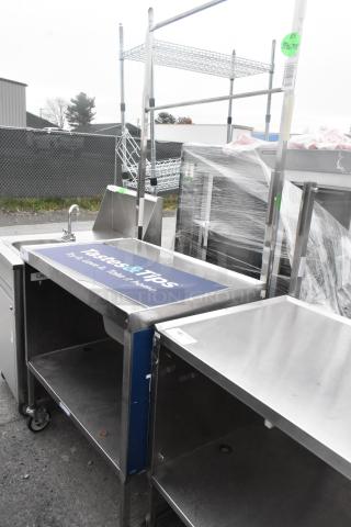 Stainless steel table with under shelf, drawer, and commercial casters. Features integrated sink and branded surface. Visible tags indicate auction ID.