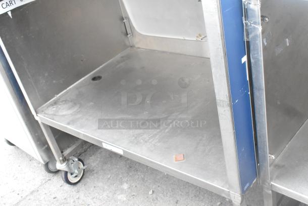 Stainless steel table with under shelf and drawer, on commercial casters. Visible wear on surfaces. Includes label for identification.