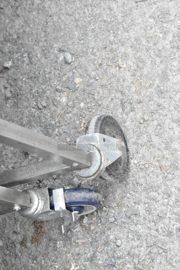 Stainless steel table on commercial casters, featuring an under shelf. Visible caster wheel on rough surface.