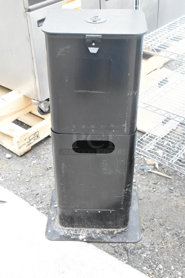 Black metal trash can, rectangular shape, minor scuffs, mounted on a base plate, suitable for outdoor or commercial use.