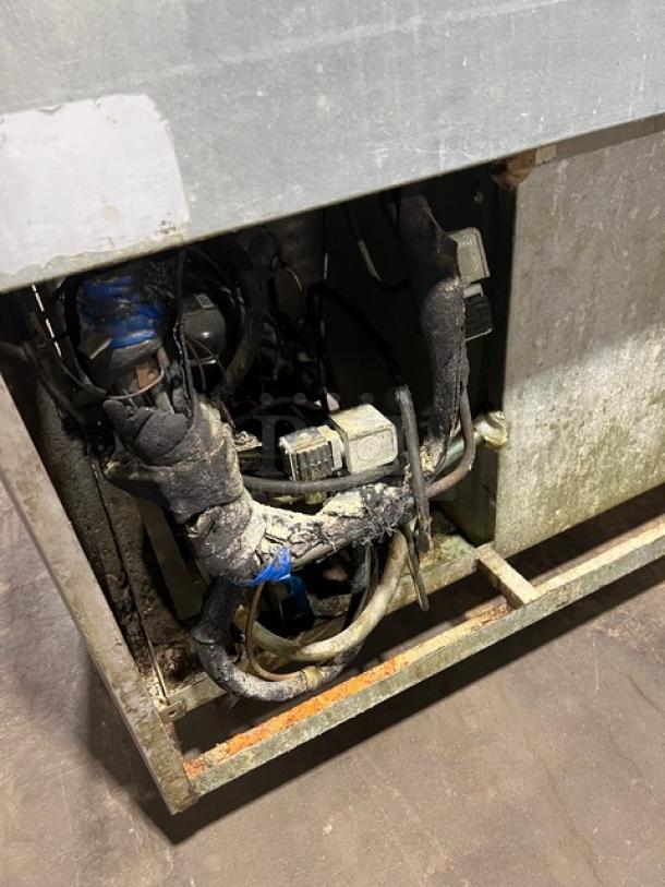 Commercial stainless steel refrigerated pizza prep table with single door, chrome-plated wire rack, and casters. Image shows exposed internal components, possibly indicating damage or repair needs.