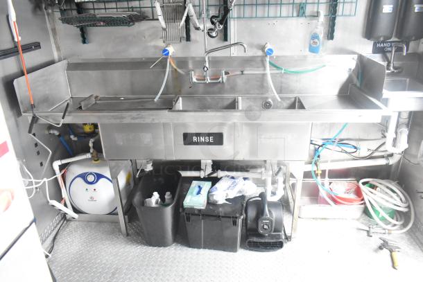 Interior of a Cruising Kitchens mobile kitchen showing a multi-compartment stainless steel sink with industrial fixtures, labeled "Rinse."