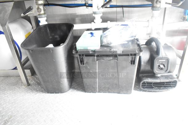 Cleaning supplies and equipment under a sink in a mobile kitchen trailer, including a trash bin, black storage box, and air mover.