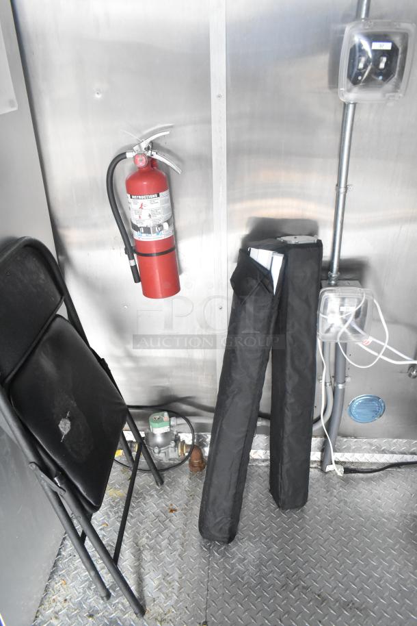 Interior of Cruising Kitchens 24' fast food trailer showing a fire extinguisher, folding chair, and utility lines on a metal floor.