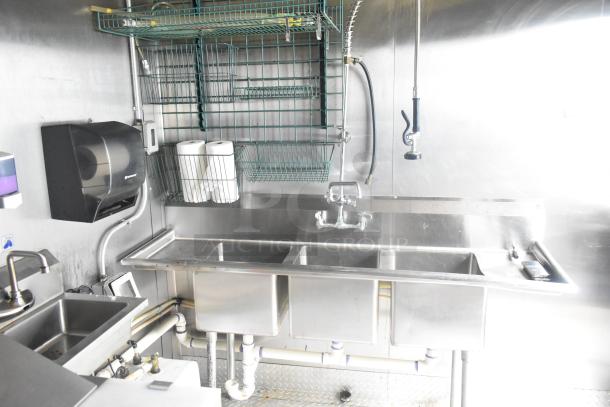 ATX 24' food trailer interior with stainless steel triple sink, wire shelving, paper towel holder, and plumbing fixtures.