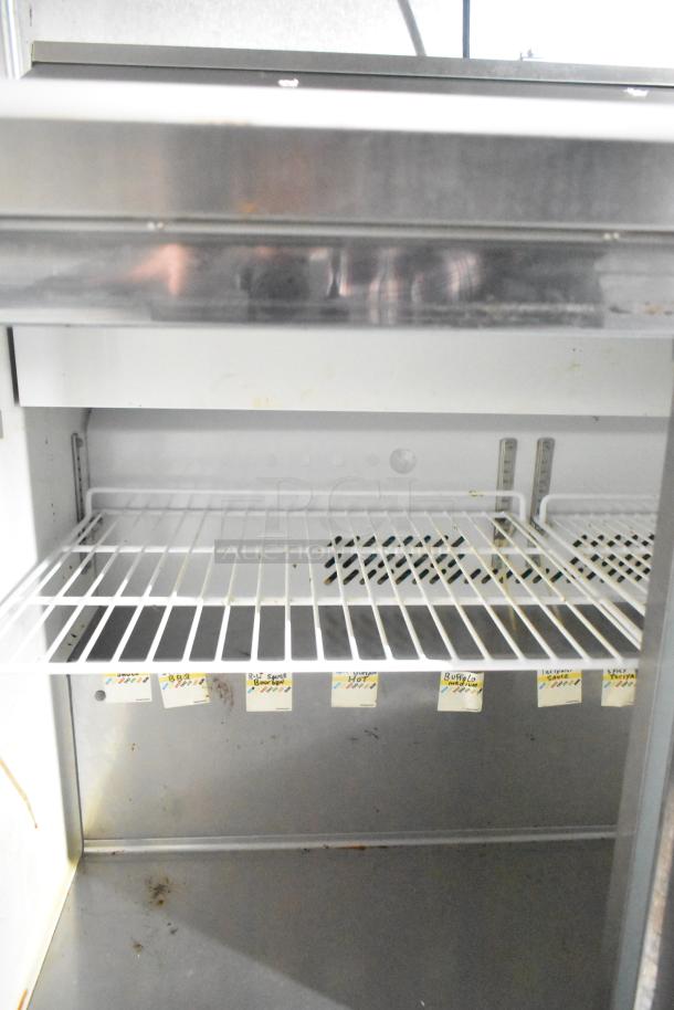 Interior view of a food trailer refrigeration unit with wire shelves. Tags labeled with various sauces are visible.
