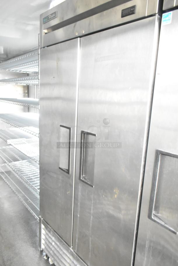 Stainless steel True reach-in refrigerator with dual doors, adjacent wire shelving units, in excellent condition.