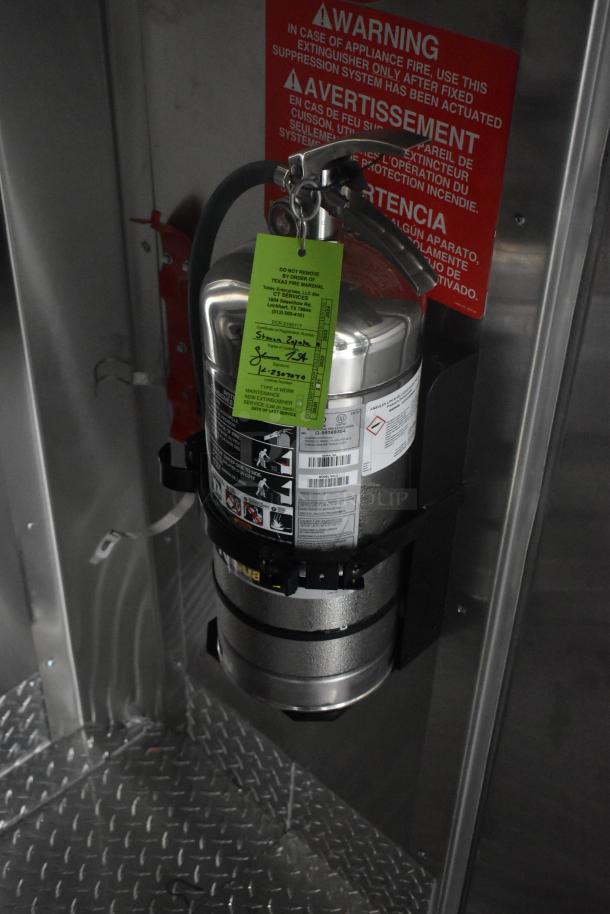 Fire extinguisher mounted inside ATX 24' food trailer kitchen with a warning sign and inspection tag visible.