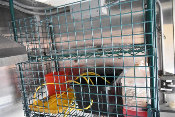 Industrial green wire shelving in a Cruising Kitchens food trailer, holding cleaning supplies and equipment, near stainless steel walls.