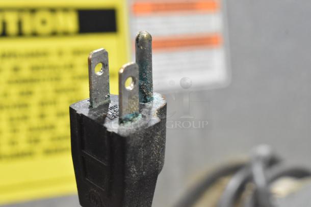 Close-up of a power plug with visible corrosion on metal prongs; caution label blurred in background.