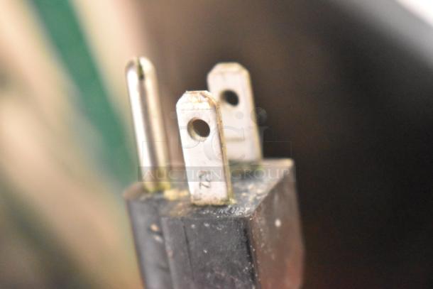 Close-up of a three-prong electrical plug, showing metal prongs and slight wear on the black casing. Plug is part of Hobart HTi-7LH26.