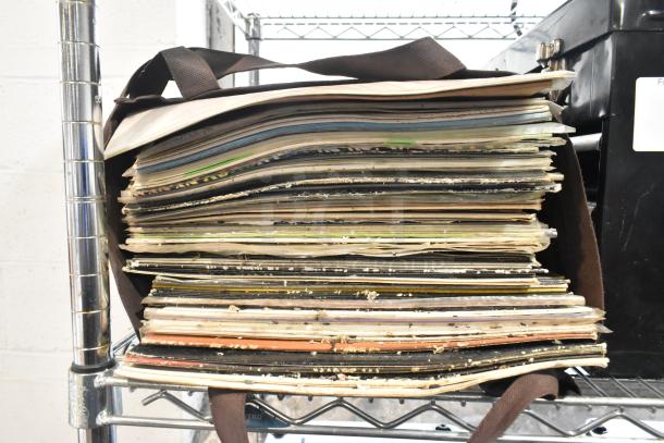 Stack of assorted vinyl records stored in a brown bag. Visible next to a black metal toolbox.