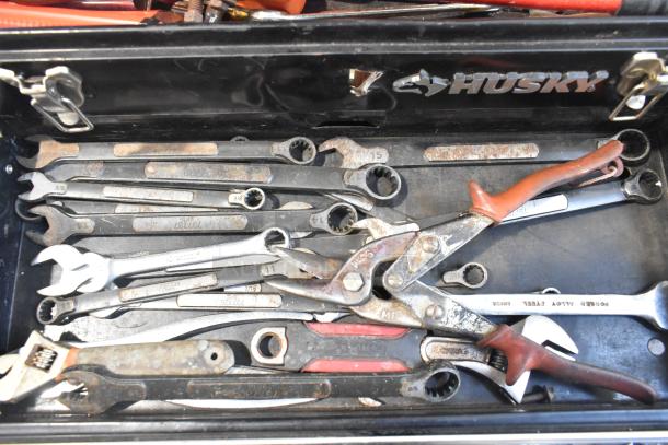 Husky black metal toolbox containing assorted tools, visible wrenches and pliers, slightly rusted, brand logo on lid.