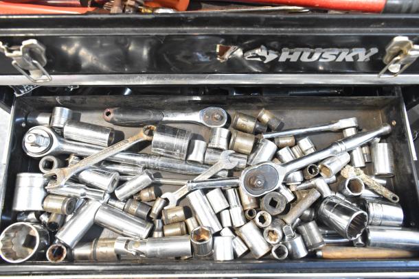 Husky black metal toolbox filled with various sockets and ratchets. Toolbox is open, showing tools in used condition.