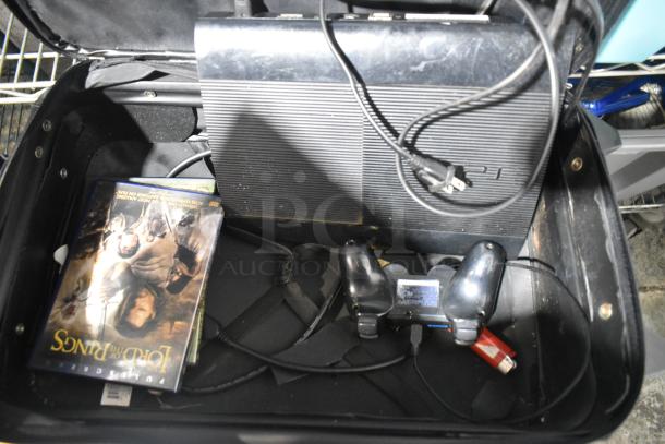A PS3 console, controller, and a "Lord of the Rings" DVD inside a black suitcase, indicating use; power cord visible.