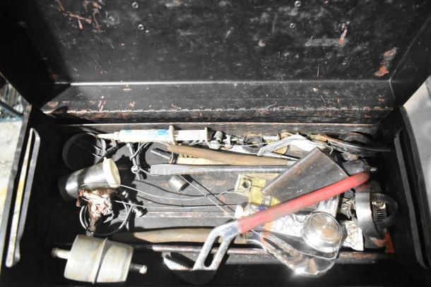 Open toolbox with various tools and items. Metal exterior with signs of rust. Contains cables, utensils, and a small brush.