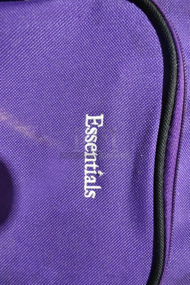 Purple Essentials luggage with white embroidered logo, part of a mixed lot including DVDs, a toolbox, and waffle makers.