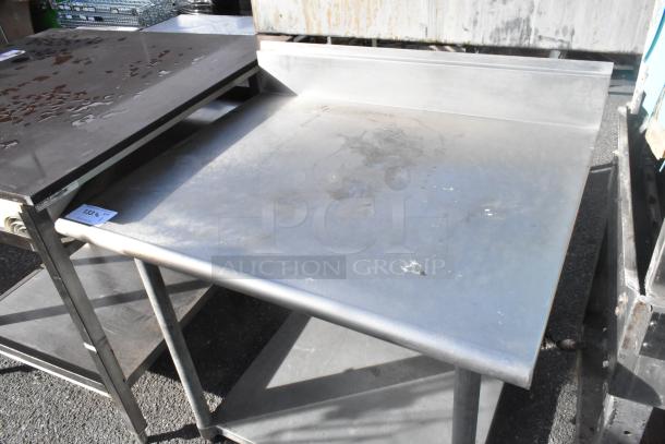 Stainless steel table with backsplash, featuring a metal undershelf. Shows surface wear. Ideal for industrial use.
