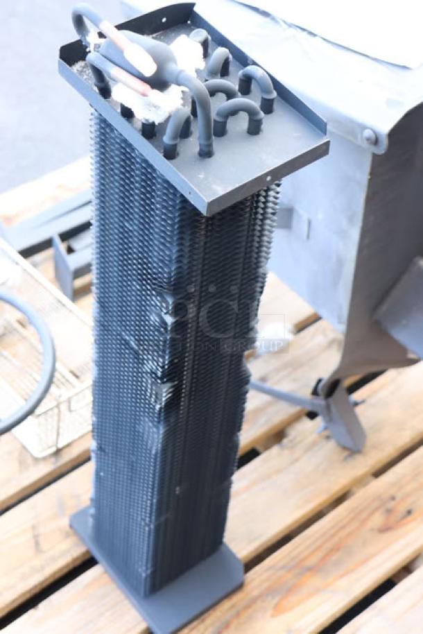 Tall black condenser coil with multiple fins and piping, standing on a wooden pallet.