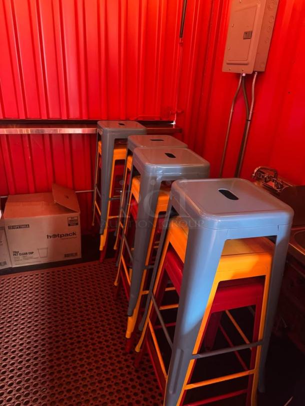 Colorful stackable metal stools inside red-walled mobile beach bar container, with visible electrical panel and cardboard box.