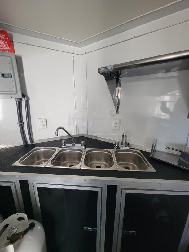 Four-compartment sink with faucets in a 2021 Covered Wagon 16' mobile food trailer's commercial kitchen. Features white walls and propane tank.