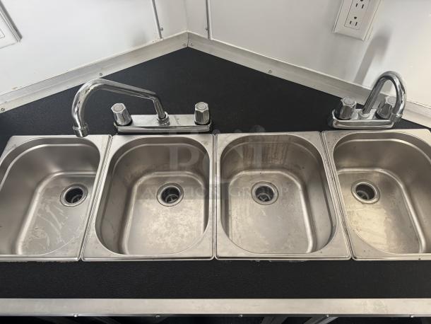 Four-compartment sink with chrome faucet in a mobile food trailer kitchen. Black countertop, outlet on wall.