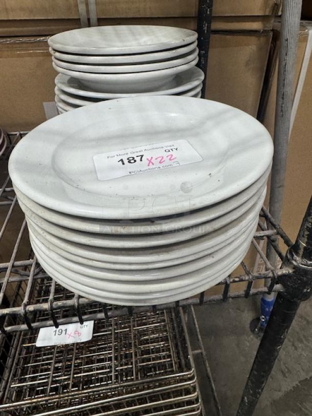 Bright white wide rim rolled edge stoneware plates stacked on a metal rack. Visible lot tag with auction details.