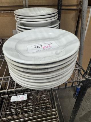 Bright white wide rim rolled edge stoneware plates stacked on a metal rack. Visible lot tag with auction details.