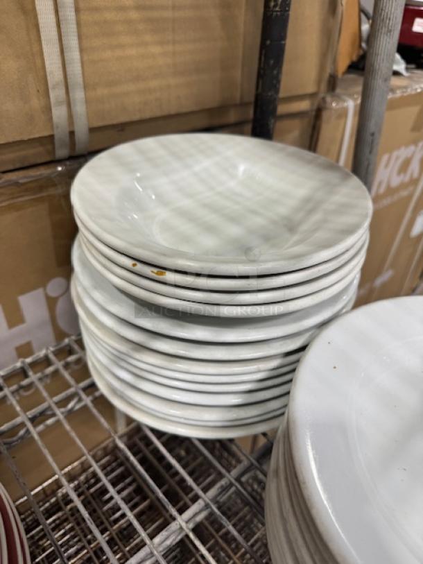 Bright white stoneware plates with wide rim and rolled edge, stacked on a shelf. Used condition, slight discolorations visible.