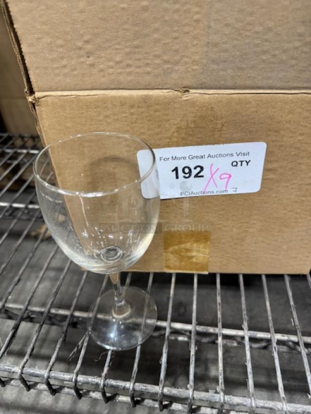 8 oz. customizable wine glass in good condition, clear glass with no visible markings, next to cardboard box on metal shelf.