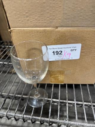 8 oz. customizable wine glass in good condition, clear glass with no visible markings, next to cardboard box on metal shelf.