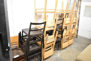 16 assorted wooden dining chairs in various shades, stacked. Good condition. Ideal for bulk purchase or event seating.