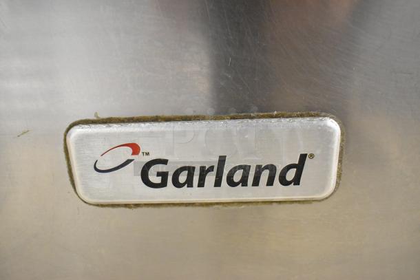 Garland stainless steel commercial range, 6 burners, gas-powered with oven and backsplash. Good condition. Garland logo visible.