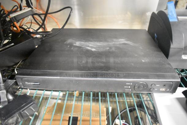 Alarm.com black electronic device with buttons and status indicators, resting on a wire shelf amid tangled cables.