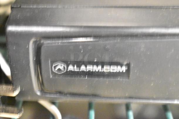 Close-up of an Alarm.com device with label. Condition appears used. Part of an 8-item auction lot.