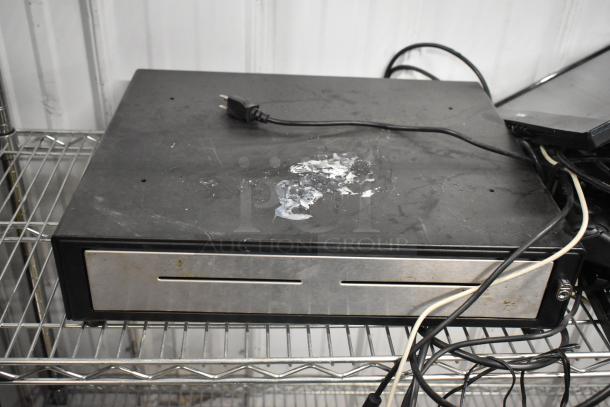 Metal cash drawer with visible wear, including scratches and adhesive residue on the top surface. Power cord attached.