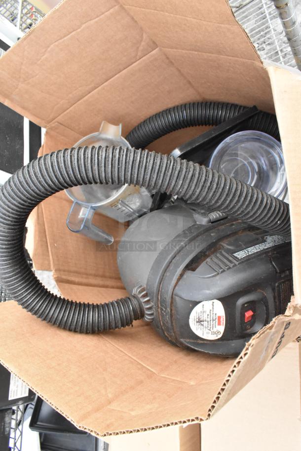 Cardboard box with vacuum hose and accessories, stored on chrome shelving unit, visible wire racks. Buyer dismantles for shipping.
