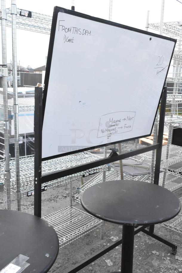 Alt text: Whiteboard on stand with writing, two round black tables, industrial setting. Signs of wear on tables.
