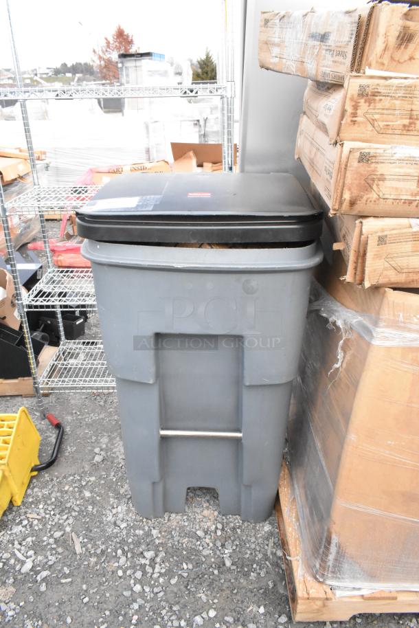 Gray poly trash can with a black lid, containing a fan and basket, located outdoors next to shelving and boxes.