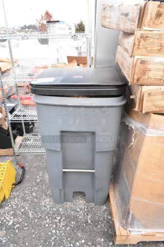 Gray poly trash can with a black lid, containing a fan and basket, located outdoors next to shelving and boxes.