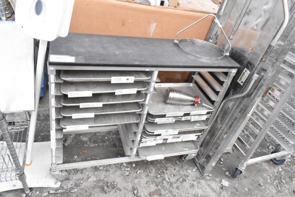 Metal commercial double pan transport rack with multiple metal baking pans, on commercial casters. Some wear visible.