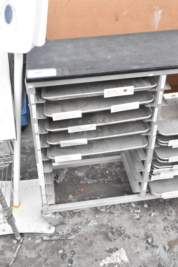 Metal commercial double pan transport rack with several metal baking pans, on commercial casters, showing visible labeling for organization.