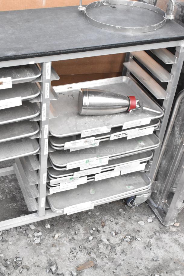 Metal commercial double pan transport rack with multiple metal baking pans, labeled for organization, on commercial casters.