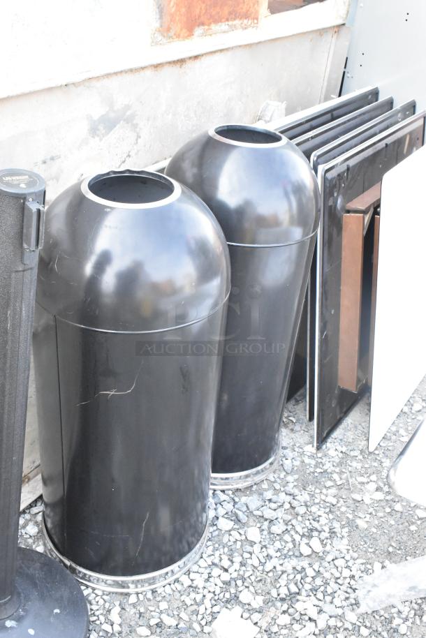 Two black metal trash can shells and one stanchion, all with minor wear, on gravel. Includes additional stanchion not shown.