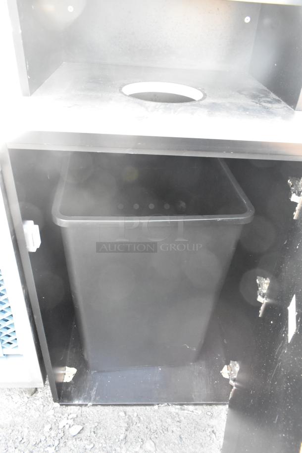 Black trash can shell with trash deposit hole, tray return, door, and poly trash can inside. Appears in used condition.