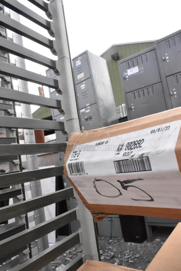 Cardboard box labeled "TTS-3 Tray Shelf" with barcode and date. Warehouse backdrop with metal shelves and lockers.