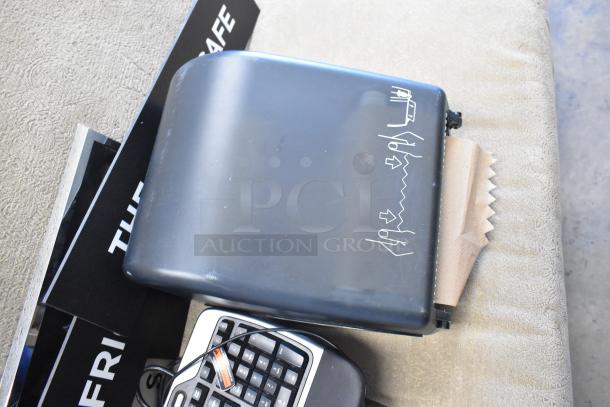 Lot of items including black paper towel dispenser with paper protruding, black keyboard, and partial signage. Mixed condition.