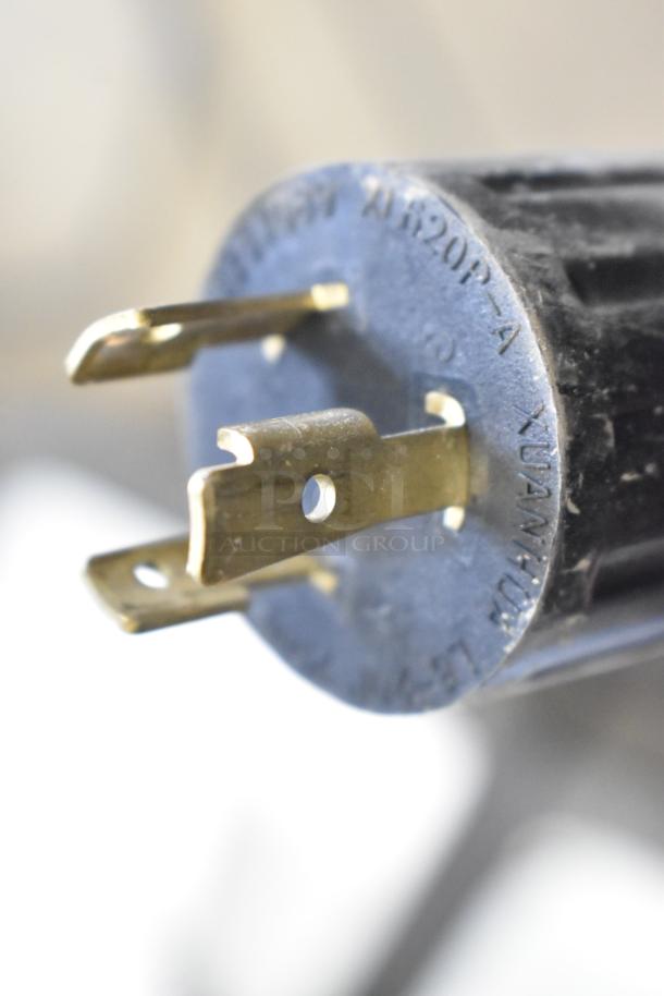 Close-up of an electrical plug with three brass prongs. The plug shows the manufacturer's details engraved around the edges.