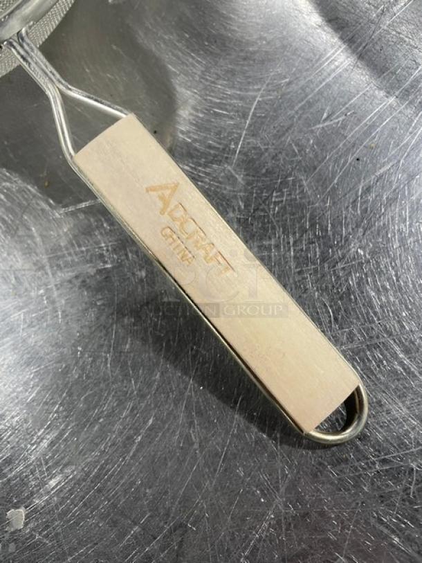 Adcraft stainless steel strainer with hardwood handle, featuring "Adcraft China" engraving. Durable, heavy-duty kitchen tool.
