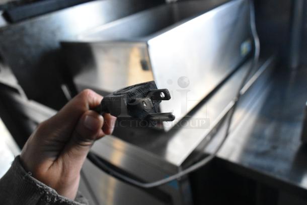 Hand holding a three-prong electrical plug in front of stainless steel kitchen equipment inside a mobile kitchen trailer.